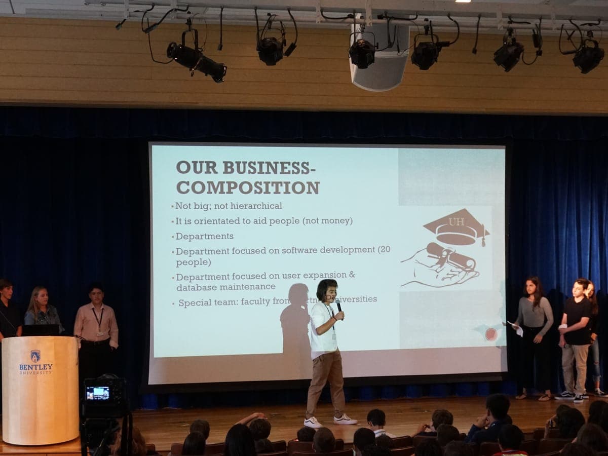 A participant alone at center stage, microphone in hand, presenting the project with the team's slide projected behind.