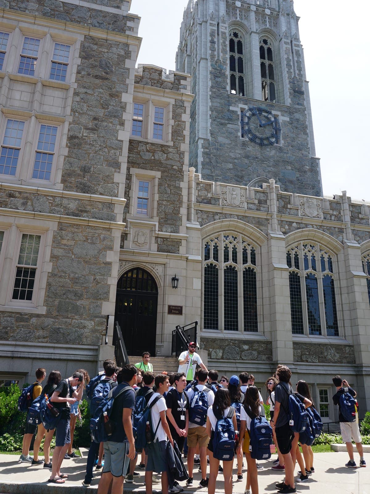 The cohort in backpacks gathered on a sunlit plaza during a visit, listening to the guide at the top of the entrance steps, campus tower behind them.