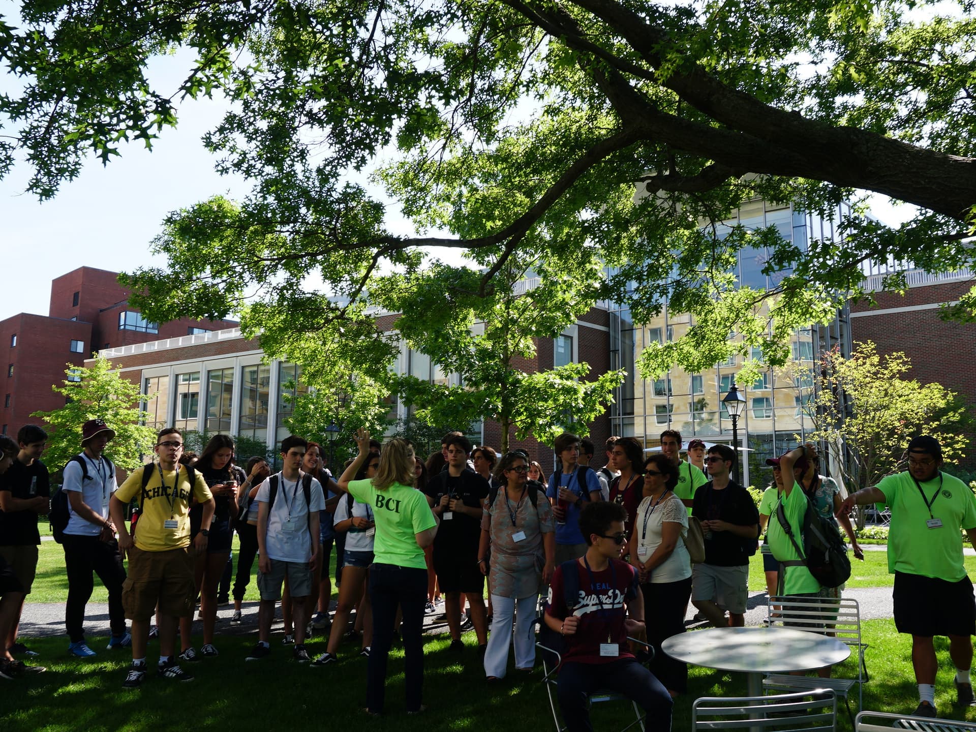 BCI mentor in the green program shirt talking with the group gathered in the shade of a tree, two more monitors alongside.