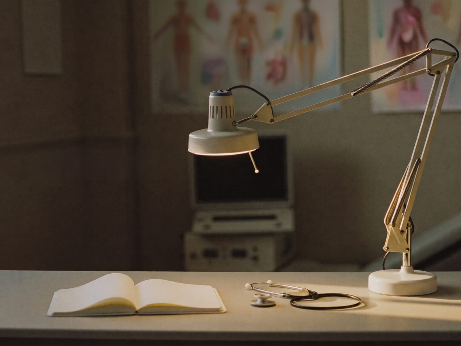 Illustrative image of a clinical desk in late afternoon light, with an examination lamp, an open blank notebook, a stethoscope and anatomy charts softly out of focus.