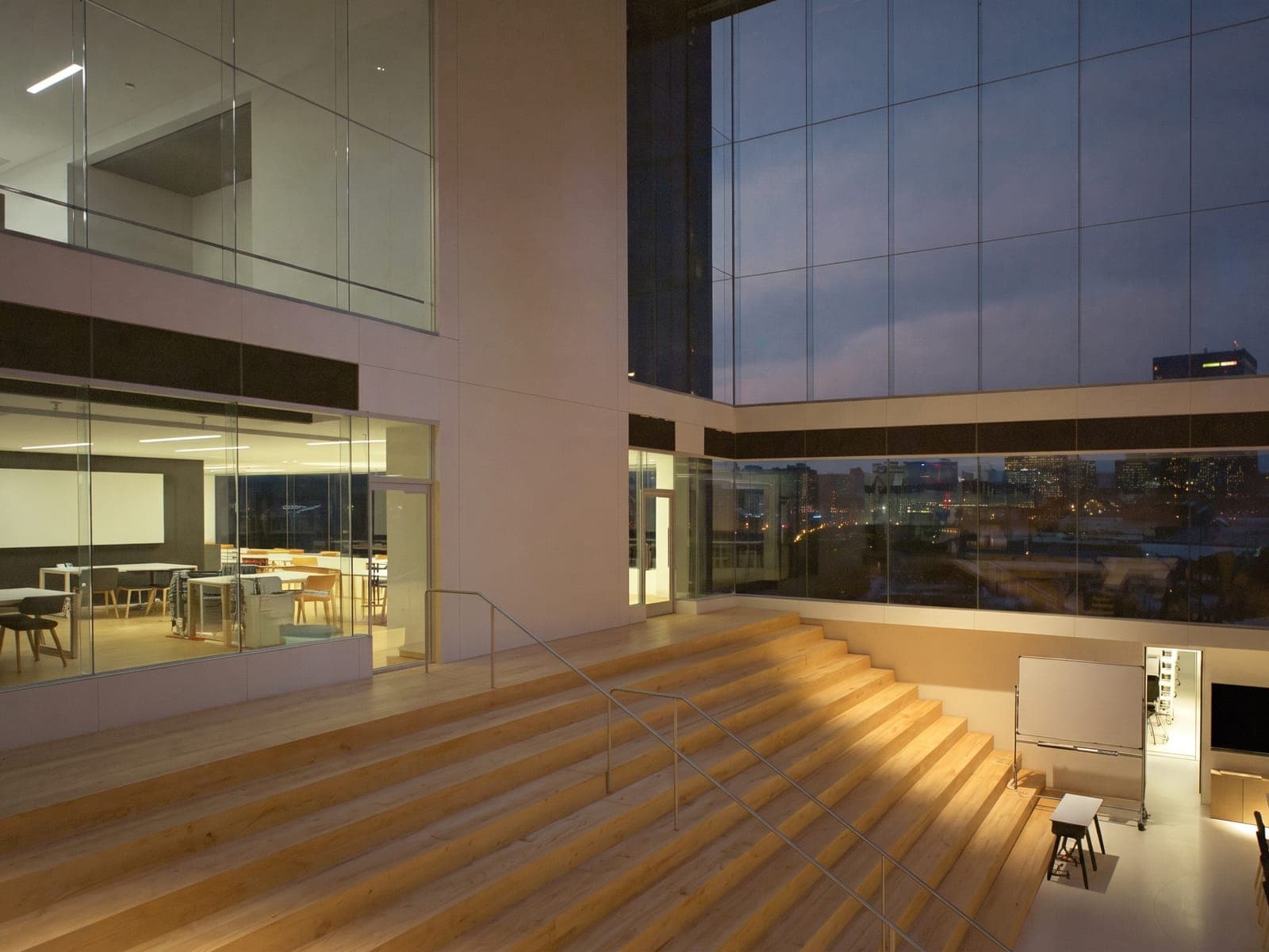 Illustrative image of a university innovation space at dusk, with tiered wooden seating, glowing glass study rooms and city lights beyond the window wall.
