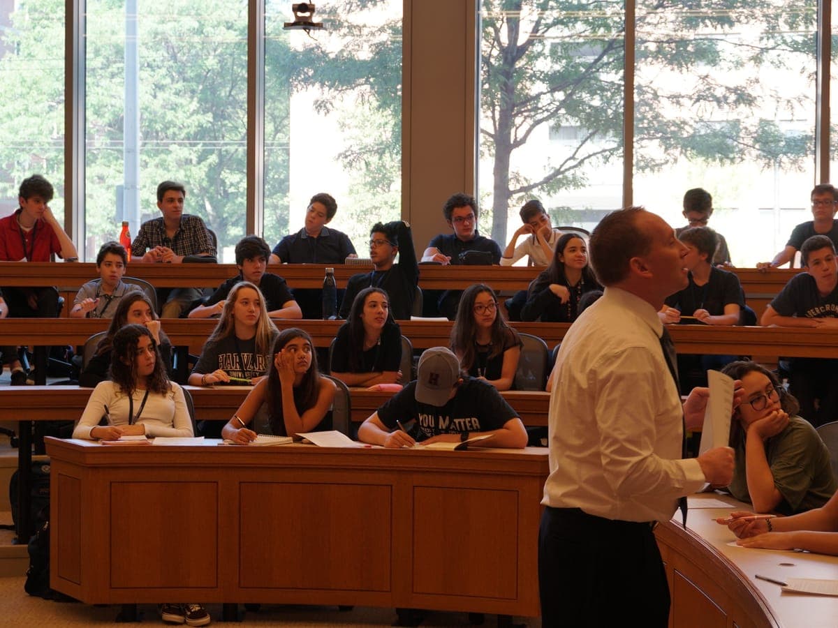 Professor in shirt and tie leading the class with the handout in hand, the group on three tiers of wooden desks, a glass wall of green trees behind them.