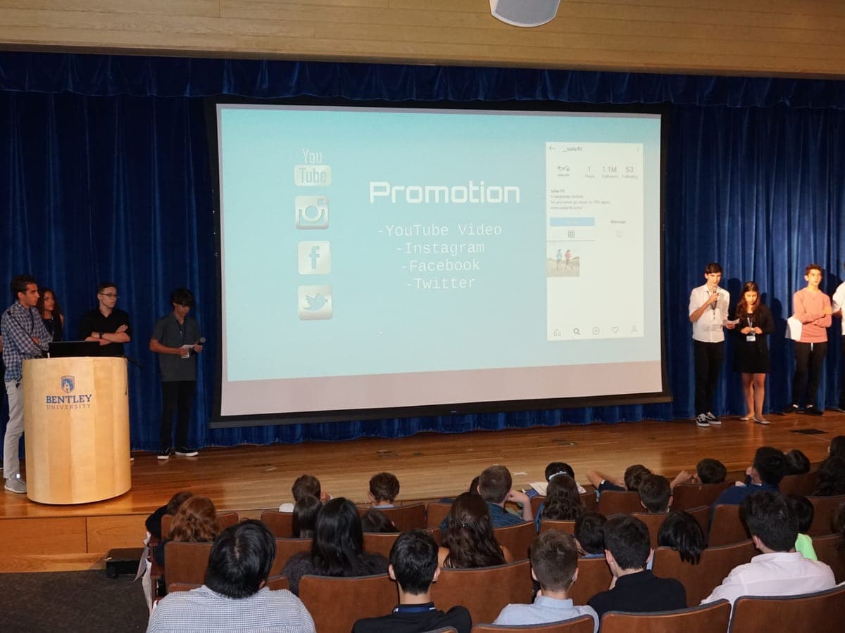Team on stage presenting their project's promotion plan on the big screen, another team waiting stage right and the audience seated in the foreground.