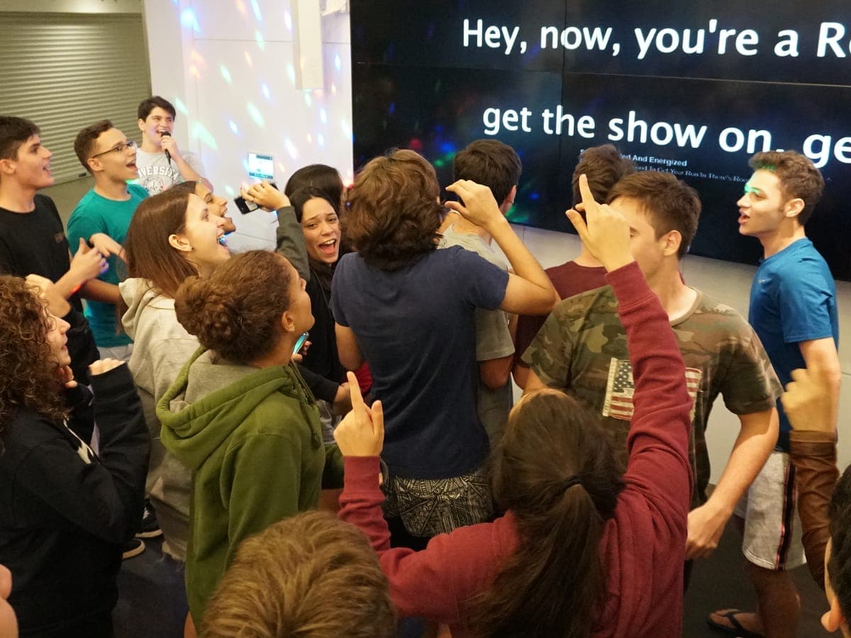 Karaoke night on campus, the group in a circle singing along under colored lights, the lyrics up on the video wall.